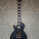 Černá elektrická kytara typu singlecut s hnědým lemováním. Na hlavě je zlaté logo Ltd a nápis ESP. Kytara má dva humbuckery se zlatými kryty, zlatý m…