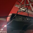 Černý aktivní reproduktor v plastovém korpusu, který je zavěšen na hliníkovém trámovém systému. Na zadním panelu jsou vidět ovládací prvky a konektor…