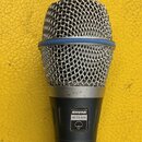 Dynamický mikrofon s kovovou síťkou a modrým kroužkem v horní části. Na těle je viditelný štítek s nápisy SHURE a BETA 87A spolu s logem značky. Povr…