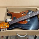 Elektrická kytara v kartonové přepravní krabici. Nástroj má černé tělo s viditelnou strukturou dřeva, hnědý pickguard a hnědý hmatník. Na hlavě kytar…