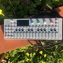 Stříbrný syntezátor s názvem OP-1 na pravém okraji. Zařízení má displej, čtyři barevné ovládací knoflíky (modrý, zelený, žlutý a červený) a bílé kláv…