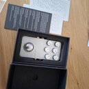 Stříbrný efektový pedál v černé krabici s přiloženým manuálem. Na pedálu je nápis XTOMP a šest bílých oválných ovládacích knoflíků. Povrch pedálu i k…