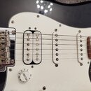 Černá elektrická kytara s bílým pickguardem. Nástroj má tři snímače, přičemž kobylní snímač je typu humbucker. Na kovových sedlích kobylníku je vidit…