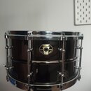 Černý malý snare drum s chromovaným kováním. Na koruse je umístěn oválný štítek s nápisy „pawpaw“, „BLACK MAGIC“ a „SNARE DRUM LIMITED“. Povrch nástr…