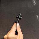 Ruka drží černý textilní popruh s kovovým připevňovacím mechanismem. Kovová část je černá a má vroubkovaný povrch pro lepší uchopení. Na popruhu ani…