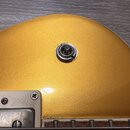 Detail zlaté kytary s kovovým vstupním jackem. Povrch nástroje má metalický les a v dolní části je vidět bílý plastový prvek s kovovým šroubem. Na fo…