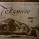 Detailní záběr na starý, zažloutlý štítek s nápisem Takamine. Na štítku je zobrazen černobílý obrázek horské krajiny a texty EF360SG, Mode oraz Made…