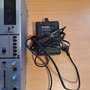 Fotografie zachycuje část stříbrného míchacího pultu s logem BEHRINGER a černý napájecí adaptér. Na adaptéru je štítek s nápisy BEHRINGER®, EURORACK…
