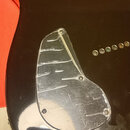 Detail korpusu tmavě hnědé elektrické kytary. Na povrchu je připevněn průhledný plastový ochranný kryt (pickguard) s viditelným opotřebením ve formě…