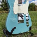 Světle modrá elektrická kytara typu Telecaster s bílým pickguardem a hnedým hmatníkem. Nástroj je umístěn v černém stojanu na trávě. Na kytare nejsou…