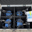 Otevřený hliníkový transportní kufr s černou pěnovou výplní obsahuje čtyři modré jednotky s připojenými kabely a svorkami. V pravé části je v bublink…