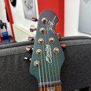 Fotografie zachycuje hlavu elektrické kytary v tmavě šedé nebo modrošedé barvě. Na povrchu je bílý nápis „Sterling by MUSIC MAN“ a stylizovaný podpis…