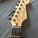 Hlava elektrické kytary z přírodního dřeva s šesti kovovými ladičkami. Na povrchu je viditelné logo Fender a nápis STRATOCASTER. Hrabednice má tmavý…