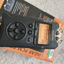 Černý digitální nahrávač TASCAM DR-40 LINEAR PCM RECORDER s displejem a ovládacími tlačítky. Zařízení leží na oranžovém originálním obalu. Na horní s…