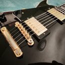 Detailní pohled na tělo černé elektrické kytary s lesklým povrchem. Nástroj má dva zlaté humbuckery, zlatý kobylník a zlatý struník. Na těle jsou umí…