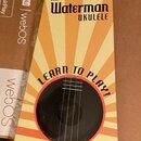 Papírové obalové pouzdro na ukulele v žlutě, oranžové a modré barvě. Na horní části je logo KALA BRAND MUSIC CO. a uprostřed nápis The Waterman UKULE…