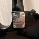 Detail zadní strany černé kytary v místě spoje krku s tělem. Je zde vidět kovový kryt připevněný čtyřmi šrouby, na kterém je vyryto logo Epiphone a t…