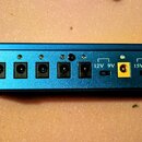 Modrý kovový napájecí adaptér s řadou konektorů. Na panelu jsou viditelné nápisy 12V 9V a 15V 9V. Zařízení obsahuje několik černých a žlutých vstupů/…