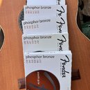 Pět bílých krabiček s nylonovými strunami značky Fender, které leží na akustické kytáře. Na každém obalu je nápis phosphor bronze a seznam kalibrů .0…
