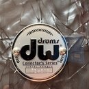 Kruhová kovová cedule s logem značky připevněná dvěma šrouby na tmavý, texturovaný povrch nástroje. Na ceduli je černý nápis drums dw, pod ním text C…