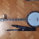Na fotografii je banjo s bílou membránou a kovovým tělem, k němuž je přiložen černý popruh a malá krabička s příslušením. Na hmatníku jsou vidět orna…