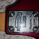Detail kovového šiltíku na červeném povrchu nástroje, připevněného čtyřmi šrouby. Na šiltíku je svisle zapsáno logo KRAMER a nápis Neptune, N.J. USA.…