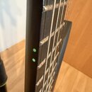 Detail krku strunového nástroje s černým hmatníkem a černým povrchem. Na hraně krku jsou vidět dvě zelené tečky sloužící jako značky poloh. Na fotce…