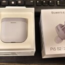 Bílé bezdrátová sluchátka v nabíjecím pouzdře umístěná v otevřené bílé krabičce. Vedle je uzavřená produktová krabice s nápisy Bowers & Wilkins a Pi5…