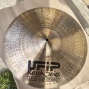 Zlatavý kovový činel s kladivkovaným povrchem, položený na hnědém kartonu. Na povrchu je černý nápis UFIP Rough Series EARCREATED CYMBALS a malá nále…