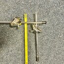 Dvě chromované kovové ramena s černými plastovými koncovkami a úchopy, pravděpodobně části stojanu. Vedle nich je položen žlutý natahovací metr. Na p…