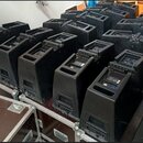 Skupina černých reproduktorů v plastovém provedení, které jsou uspořádány v řadách. Některé z nich stojí na hnědých transportních kufrech s kovovými…