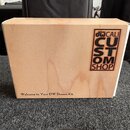 Dřevěná krabice světlé barvy s viditnou strukturou dřeva. Na pravém boku je černý nápis „drums DW CALI CUSTOM SHOP“, kde slovo „CUSTOM“ je napsáno sv…