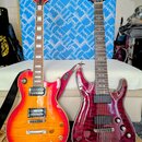 Dvě elektrické kytary opřené o bílý gauč. Levá kytara má žlutě-oranžovo-červený přechodový lak, dva humbuckery a na hlavě nápis "Squier". Pravá kytar…