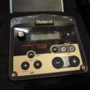 Černý trigger modul značky Roland s modelovým označením TRIGGER MODULE TM-2. Přední panel je kombinací černého plastu a zlatavé plochy s několika ovl…