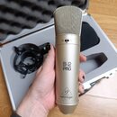 Zlatý kondenzátorový mikrofon s mřížkou v horní části. Na těle je černý nápis B-2 PRO a níže logo s textem behringer. Mikrofon je v ruce, v pozadí je…