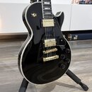 Černá elektrická kytara s tvarem singlecut a bílým lemováním hran. Nástroj má zlatý hardware, včetně dvou humbuckerů a mostu. Na horní části těla je…
