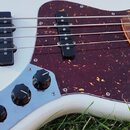 Bílá elektrická basová kytara s tmavě červeným vzorovaným pickguardem. Nástroj má dva černé snímače a tři černé ovládací knoflíky na chromovaném pane…