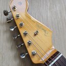 Hlava elektrické kytary z přírodního dřeva s nápisem Fender a STRATOCASTER. Na hlavě jsou vidět čtyři ladicí mechanismy z chromovaného kovu a nápis W…