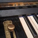 Detail klaviatury černého pianina s viditelnými bílými a černými klávesami. Na černém povrchu je přišroubovaná mosazná kovová lišta s knoflíkem a náp…