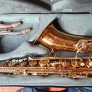 V otevřeném kufru značky Bam leží zlatý saxofon a dva zakřivené hrdly s korkem. Nástroj má lesklý povrch a je vybaven řadou klapek a mechanismů. Na s…