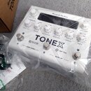 Bílý kytarový procesor v ochranné plastové fólii. Na horní straně je nápis AmpliTube, uprostřed velký text TONEX a dále nápisy MACHINE MODELING a VIR…