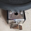 Detailní záběr na spodní část černého elektronického bubnu s nápisem Roland. Nástroj je upevněn do chromovaného kovového držáku s šroubem a motýlkový…