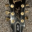 Černá hlava elektrické kytary se zlatými ladičkami. Na povrchu jsou zlaté nápisy Gibson, MODEL Les Paul a Studio. Povrch vykazuje drobné škrábance a…
