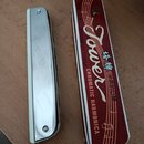 Na fotografii je chromovaná chromatická harmonika a její červené kovové pouzdro. Na nástroji je vyražen nápis TOWER, CHROMATIC HARMONICA a MADE IN CH…