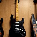 Černá elektrická kytara s hmatníkem ze světlého dřeva. Na hlavě je viditelné logo Fender. Kytara má tři snímače (dvě humbuckery a jeden jednosnímač),…