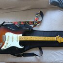 Elektrická kytara s tělem v provedení sunburst, bílým pickguardem a světlou hřednou. Na hlavě kytary je viditelné logo Fender. Kytara leží v černém s…