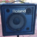 Černý kombozesilovač s povrchem z textilie a kovovou ochrannou mřížkou. Na mřížce je nápis Roland a v dolní části logo KC s doplňkem KEYBOARD AMPLIFI…