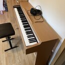 Digitální piano v dřevěném korpusu s přirozenou barvou světlého dřeva. Na klávesnici leží černá sluchátka s hlavovým obloukem a kabelem. K nástroji p…