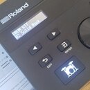 Černý elektronický modul značky Roland s nápisem V-Drums. Na displeji je zobrazen text DRUM KIT 02 Solid Rock. Modul obsahuje ovládací tlačítka ENTER…