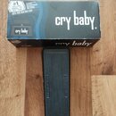 Na fotografii je černý wah pedál s gumovou texturovanou povrchovou vrstvou, umístěný před svou originální krabicí. Na krabici je bílý nápis cry baby.…
